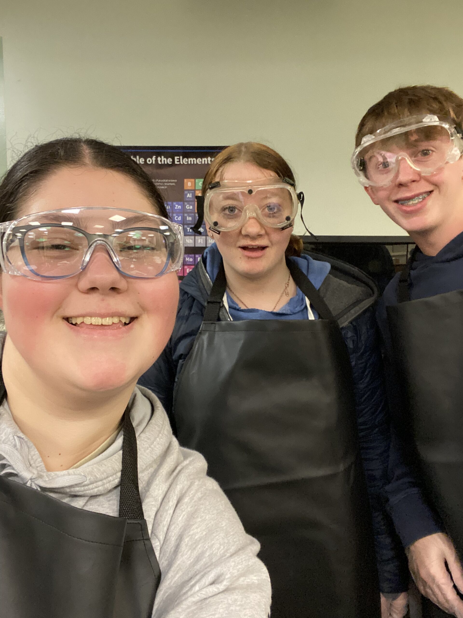 Three Grace students wearing safety goggles and aprons in a science classroom